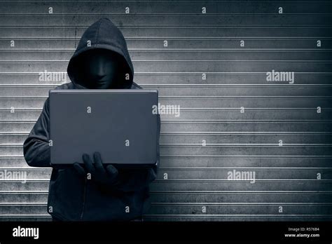 Hooded Hacker With Mask Holding Laptop While Typing Stock Photo Alamy