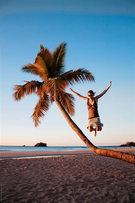 Woman Jumping Into The Sand From A Palm Tree On The Beach At Sunset