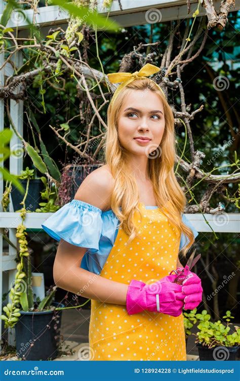 Belle Femme Blonde Songeuse Dans Le Tablier Et Les Gants En Caoutchouc Tenant Des Ciseaux Et