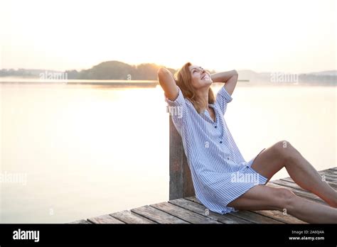Happy Mature Woman Sitting On Jetty At A Lake At Sunrise Stock Photo