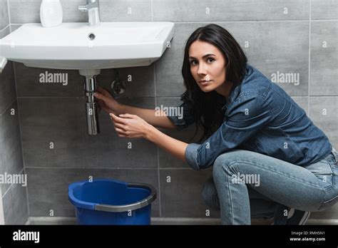 Charming Brunette Girl In Denim Shirt Repairing Pipe In Bathroom Stock