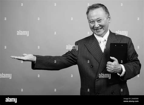 Studio Shot Of Mature Asian Businessman Holding Clipboard Against Gray