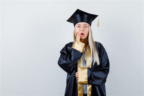 졸업 가운과 입 근처에 한 손을 넣어 모자에 금발 소녀 Bellyok에 다른 손을 잡고 무료 사진