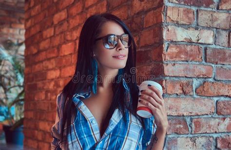 A Happy Brunette In Flannel Shirt And Sunglasses Holds A Cup Of Coffee Posing Against A Brick