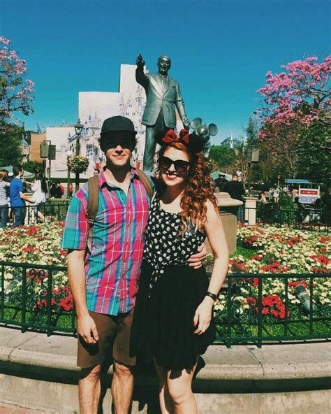 Two People Standing Next To Each Other In Front Of A Statue And Flowerbeds