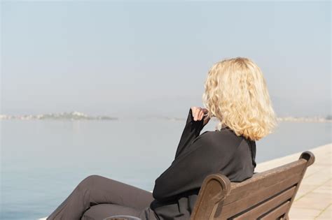 Schöne erwachsene blonde Frau sitzt auf einer Bank und blickt auf das Meer und den blauen Himmel