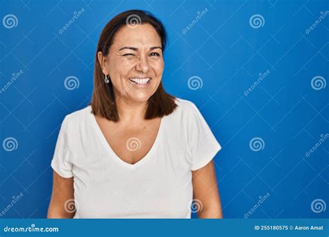 Hispanic Mature Woman Standing Over Blue Background Winking Looking At
