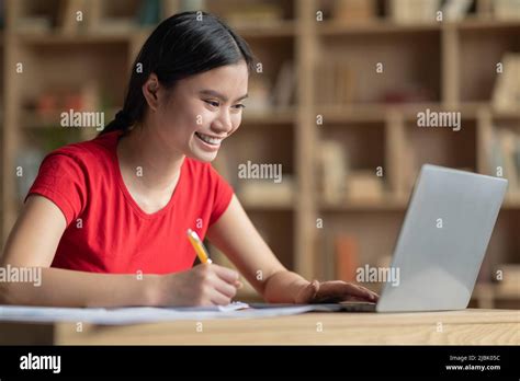 Smiling Happy Teenage Japanese Girl Doing Homework At Table With Laptop