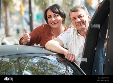 Happy Mature Couple Standing Near Twizy Electric Outside And Smiling