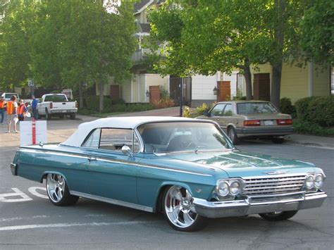 An Old Car Is Parked On The Side Of The Road In Front Of Some People