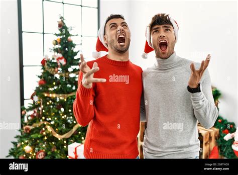 Joven pareja gay de pie por el árbol de Navidad con sombrero loco y loco gritar y gritando con