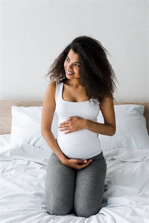 Curly African American Pregnant Woman Touching Belly While Sitting On