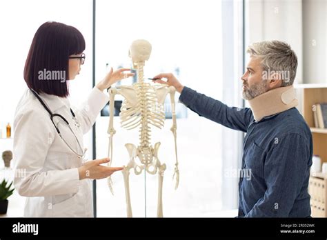 Focused Brunette With Stethoscope Indicating On Human Skeleton While Mature Male In Neck Brace