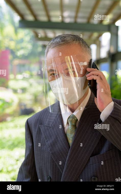 Mature Japanese Businessman With Mask And Face Shield Talking On The
