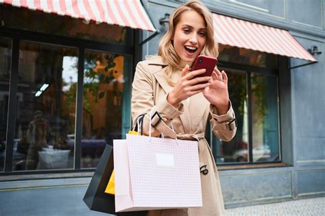 Jolie fille blonde excitée en trench coat élégant avec des sacs à provisions utilisant