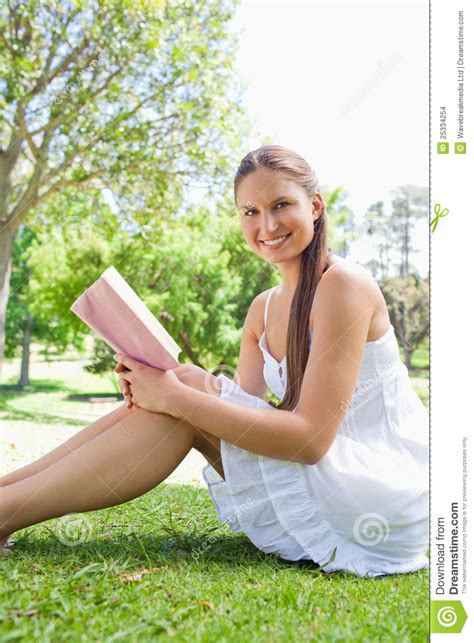 Side View Of A Smiling Woman Sitting On The Grass With A Book Stock