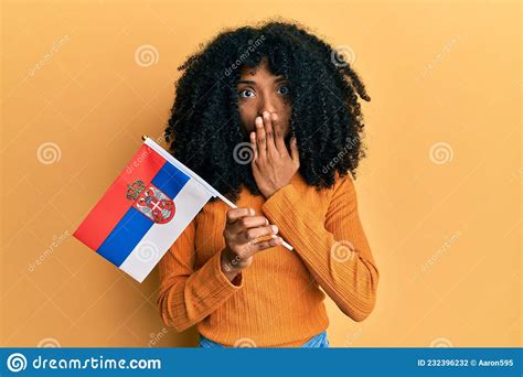 African American Woman With Afro Hair Holding Serbia Flag Covering Mouth With Hand Shocked And