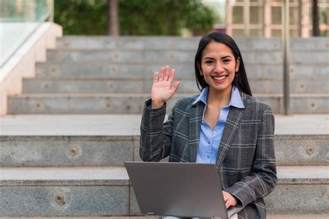 Joven empresaria latina sentada en las escaleras con la laptop saludando y mirando a la cámara