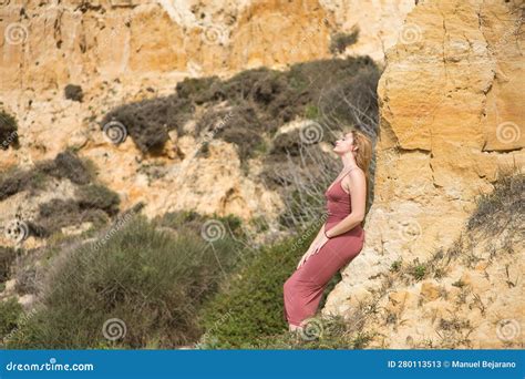 Young Beautiful Blonde Woman In A Pink Dress Sitting On A Stone Wall