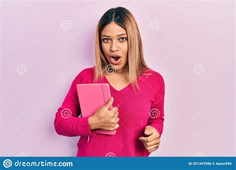 Beautiful Hispanic Woman Holding Book Scared And Amazed With Open Mouth