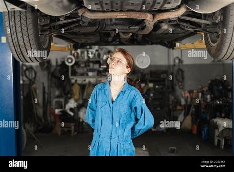 Auto Workshop And A Woman Mechanic With Glasses Stands Under The Car In