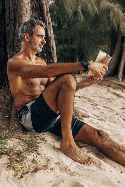 Shirtless Mature Man Reading Book While Sitting Near Tree Trunk At