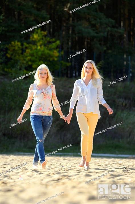 Two Happy Young Blond Women Walking Hand In Hand On Sandy Beach Stock