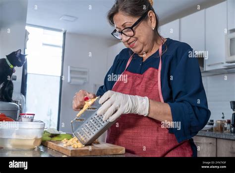 Senior Adult Latina Woman Grating Green Onion With Grater In Kitchen While Black Cat Watches Her