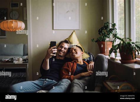 Smiling Gay Couple Sharing Smart Phone While Sitting Near Window At