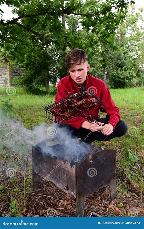 Young Man Roasts Meat Barbecue On Coals On Grill In Nature He S