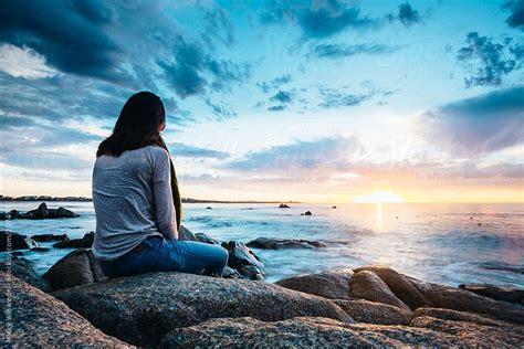 A Woman Sitting On Top Of A Rock Near The Ocean Watching The Sun Go Down