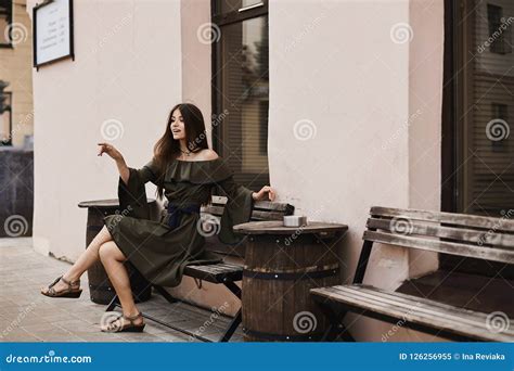 Beautiful And Fashionable Brunette Model Girl In Stylish Dress With Naked Shoulders Sits On A