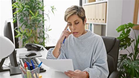 A Thoughtful Hispanic Woman With Short Blonde Hair Reviews Documents In