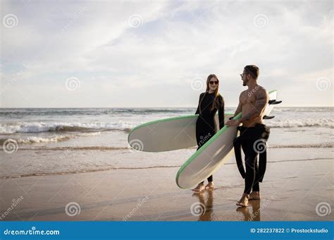 Young Couple Of Surfers Walk With Surfboards On The Beach At Sunset