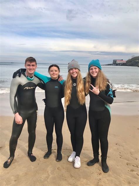 Four People In Wetsuits Standing On The Beach