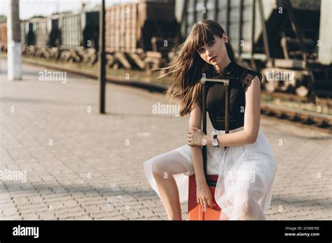 Female Brunette Traveler With Red Suitcase In White Skirt Waiting For A