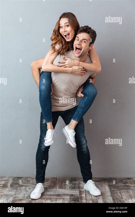 Full Length Portrait Of A Happy Man Carrying Girlfriend On His Back And Having Fun Over Gray