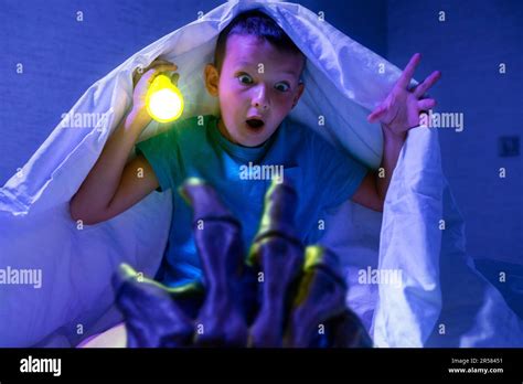 Scared Boy With Flashlight Hides Under Blanket From The Hand Of Scary