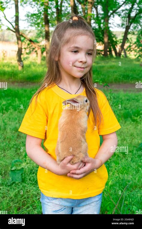 Little Cute Girl Plays With A Real Ginger Rabbit On A Background Of Green Plants Summer Outdoor