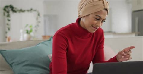 Video Of Happy Biracial Woman In Hijab Sitting On Sofa And Using Laptop