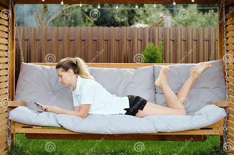 Woman Lying On Her Stomach On A Cozy Hanging Bench With A Smartphone