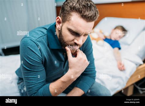 Portrait Of Thoughtful Dad Sitting Near Sick Son In Hospital Bed Stock