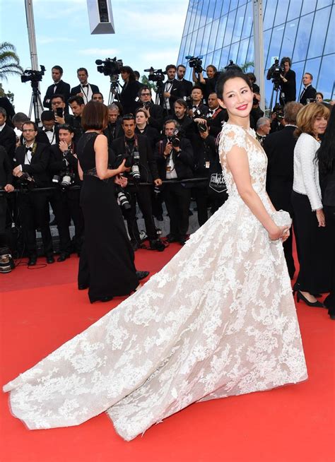 A Woman In A White Dress Standing On A Red Carpet Next To Photographers