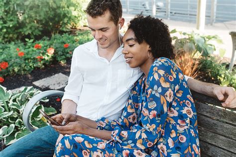 A Young Couple Sitting On A Park Bench Listening To Music By Stocksy