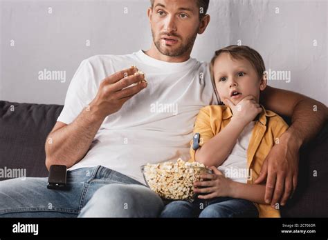 Concentrated Father And Son Watching Tv And Eating Popcorn On Sofa At