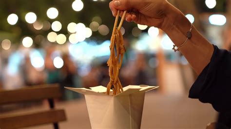 Close up of feminine hand using chopsticks to pick up noodles from a chinese take out box at an ...