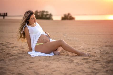 Beautiful Young Pregnant Woman Sitting On Sand Touching Her Belly And Enjoying Pregnancy On The