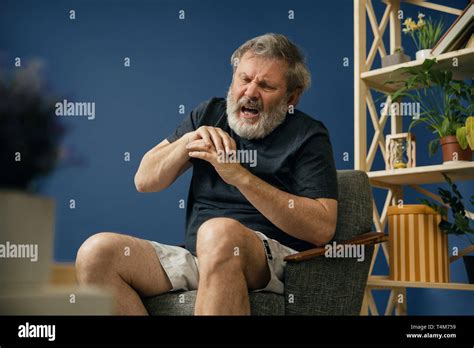 No Control Of Body Old Bearded Man Sitting On The Chair And Suffering From Pain In Hands On