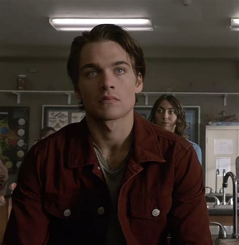 A Man In A Red Jacket Sitting At A Table With Two Other People Standing