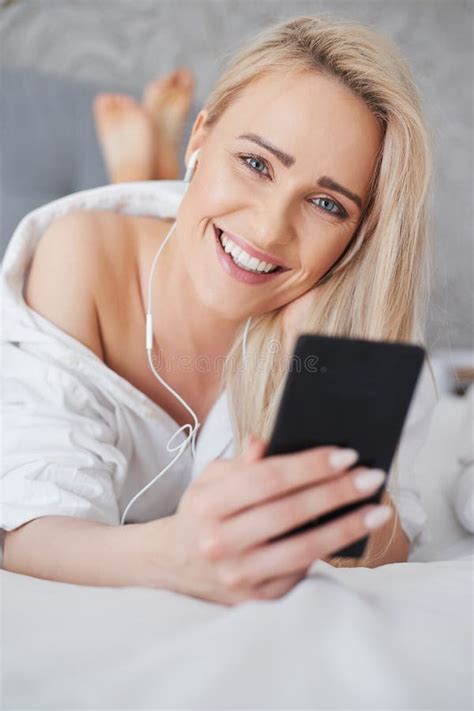 Belle Souriante Femme Blonde Se Situant Dans Le Lit Blanc Et L Aide D Un Smartphone Photo